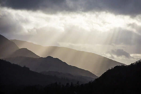 Sun rays shining through clouds onto Loughrigg Fell and hills of the Lake District National Park, in evening. Ambleside, Cumbria, England, UK. April 2020