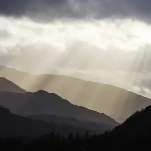 Sun rays shining through clouds onto Loughrigg Fell and hills of the Lake District National Park, in evening. Ambleside, Cumbria, England, UK. April 2020 Sun rays shining through clouds onto Loughrigg Fell and hills of the Lake District National Park, in evening. Ambleside, Cumbria, England, UK. April 2020