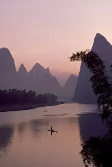 Man using pole to propel bamboo raft on Lijang / Li River, Karst peaks beyond. At dawn